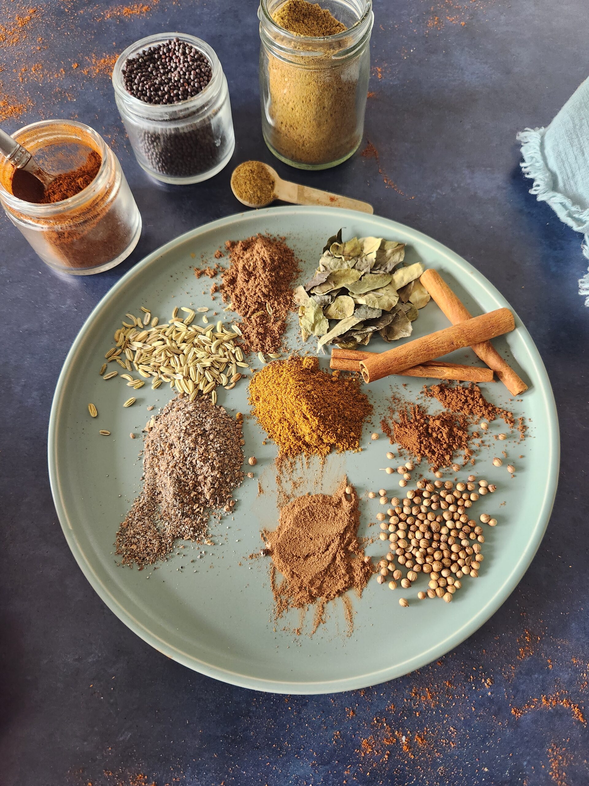 Various Indian spices on a round plate surrounded by open jars of other spices. Spices on plate include curry powder, garam masala, mustard seed, fenugreek seed, cinnamon sticks, curry leaves, and cloves