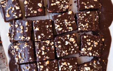 large rectangular chocolate cake on white parchment paper that has been cut into many small square slices. wooden handled knife on left hand side of photo