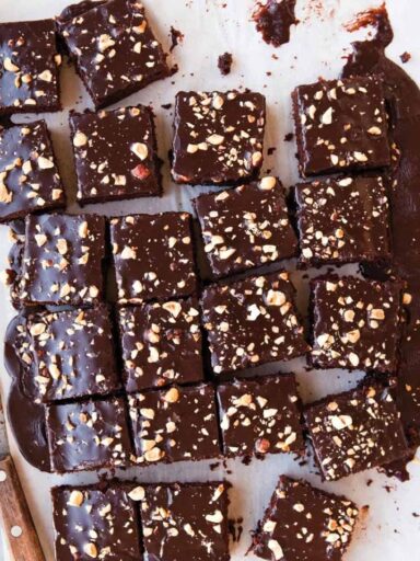 large rectangular chocolate cake on white parchment paper that has been cut into many small square slices. wooden handled knife on left hand side of photo