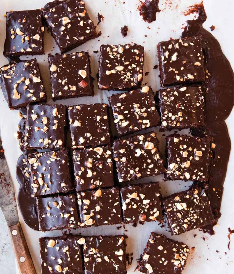 large rectangular chocolate cake on white parchment paper that has been cut into many small square slices. wooden handled knife on left hand side of photo