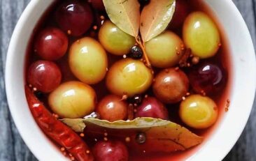 Dark Grey wood background with a white bowl filled with red and green grapes in a pickling liquid with whole spices and bay leaves.