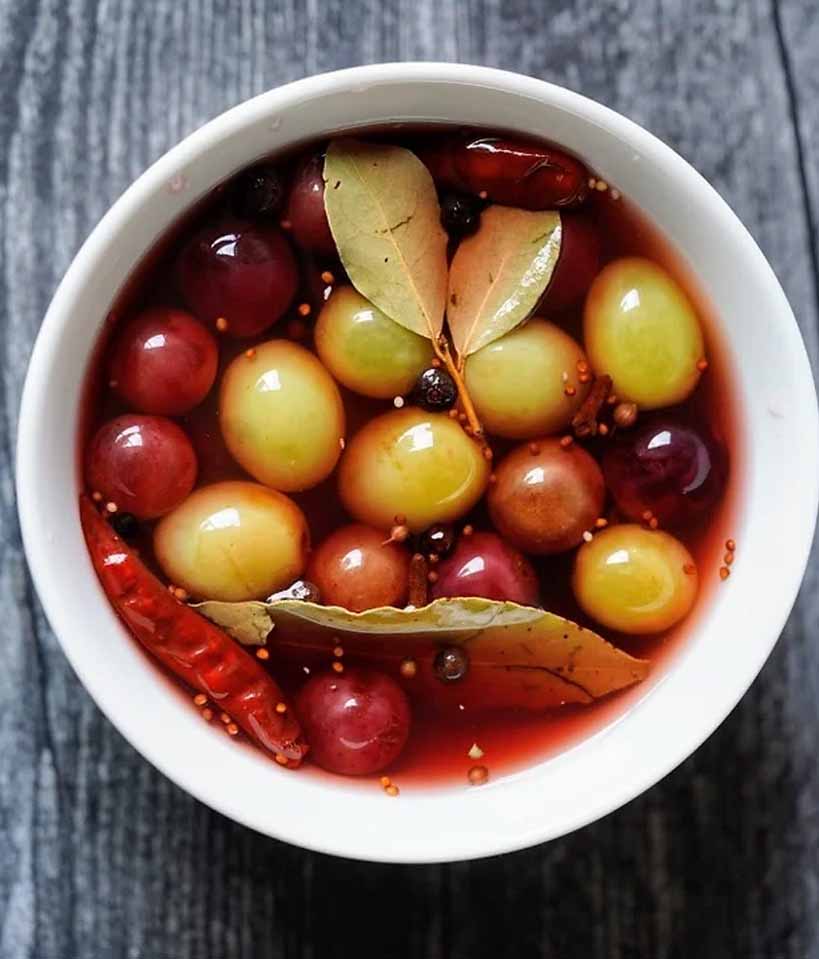 Dark Grey wood background with a white bowl filled with red and green grapes in a pickling liquid with whole spices and bay leaves.