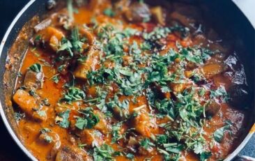 Dark blue background. Large skillet with chicken curry and fresh green herbs sprinkled on top.