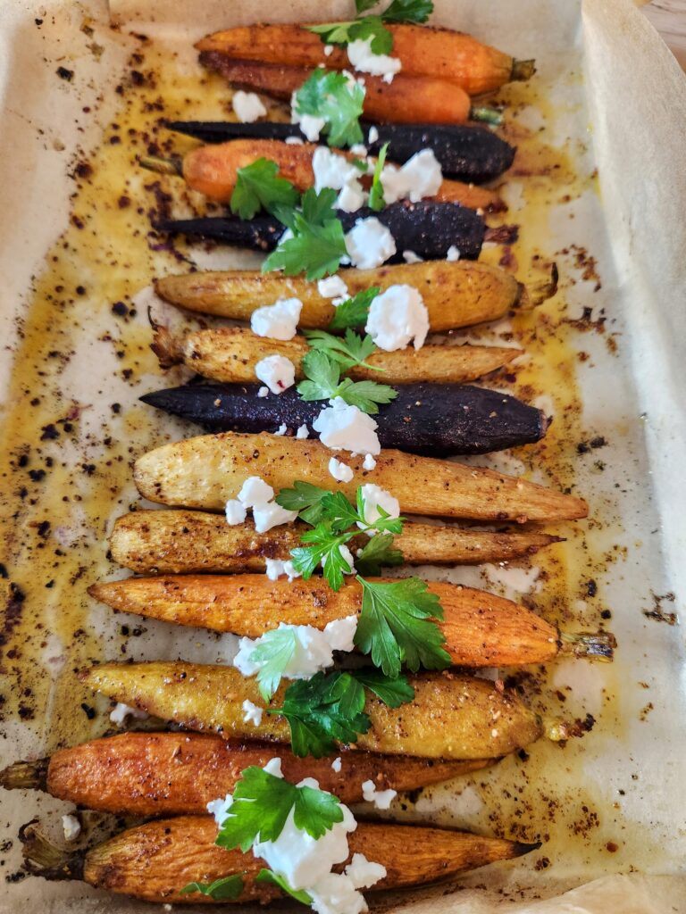 Baking sheet of multi-colored roasted carrots spiced with baharat and garnished with parsley and feta cheese.