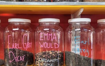 Red wall with three shelves of gallon sized glass jars of loose leaf teas.