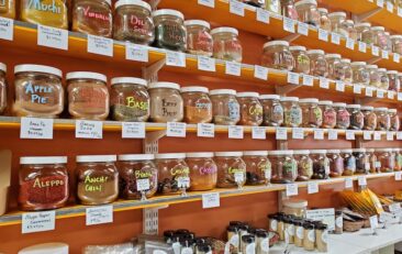 Orange wall with 6 shelves filled with hundreds of large jars of spices, herbs, salts, peppers, and chiles. Lower bottom of image shows a white counter with many small jars of spices.