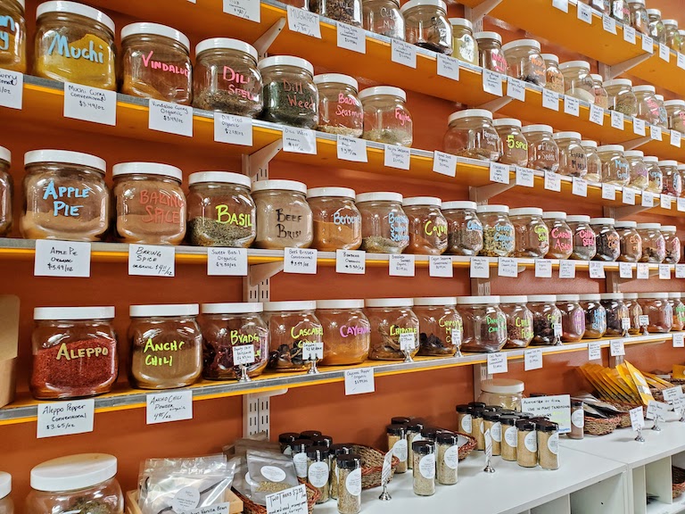 Orange wall with 6 shelves filled with hundreds of large jars of spices, herbs, salts, peppers, and chiles. Lower bottom of image shows a white counter with many small jars of spices.