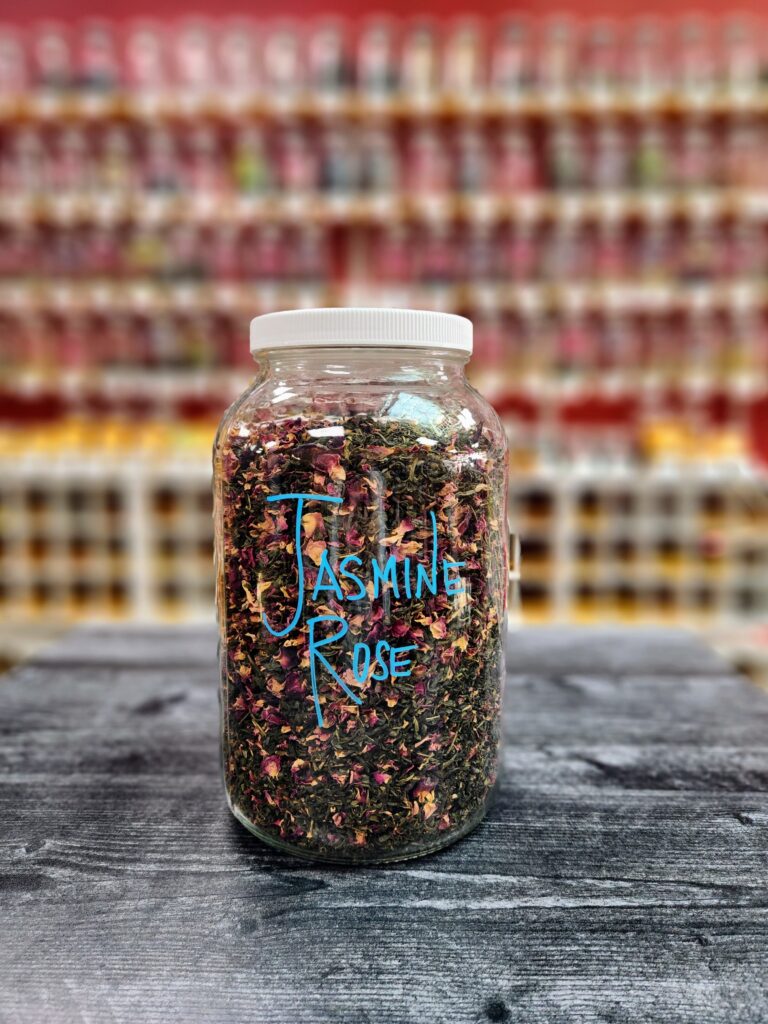 A large glass gallon jar filled with a colorful tea with the label Jasmine Rose. Wall covered with shelves of large glass gallon jars filled with different teas in the background.