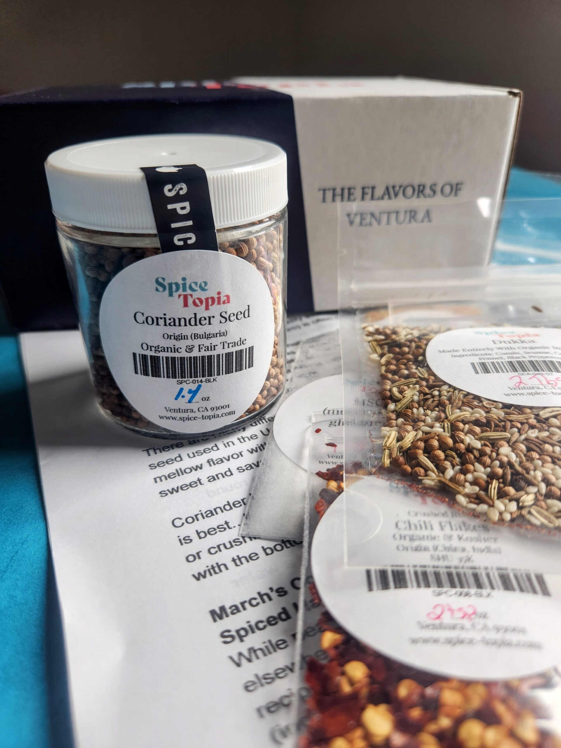 A jar of coriander in front of a SpiceTopia box with small packages of other spices scattered about.