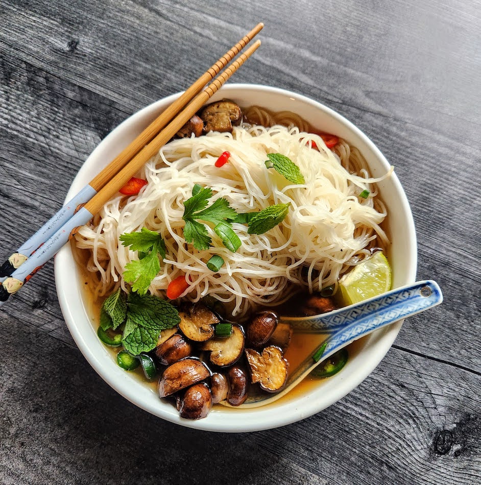 Bowl of pho soup with rice noodles, mushrooms, chilis, lime, and herbs. Bowl has a soup spoon and chopsticks